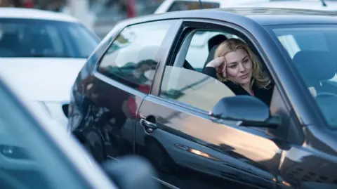 Getty Images Bored woman with head on her hand seen behind the steering wheel of her car with the window down, sitting in traffic