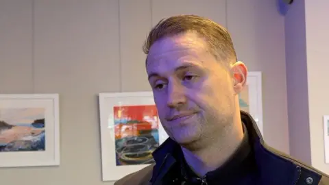 Ben Cairney is wearing a black jacket and jumper. He has short brown hair. 