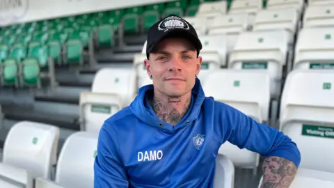 A man wearing a blue hoodie sits on a white plastic chair at what appears to be a sport ground. He is wearing a black cap and is looking into the camera. The rows of seating behind him are white and green.