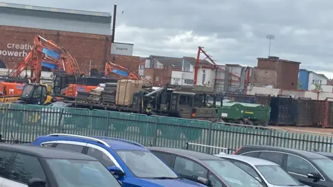 Parts of cars are in the foreground at the bottom. Behind them on the photo are railings and behind those is a building on the left and other buildings in the distance on the right.