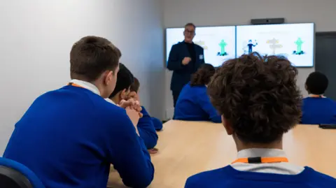 Ahead Partnership Several students wearing blue school uniform sit around a table with their backs to camera. They're listening to a man who is stood in front of a screen presenting a workshop to them.