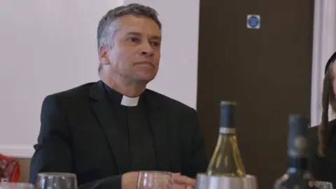 A screengrab of David Tudor, dressed in a priest's clerical collar and a dark jacket.

