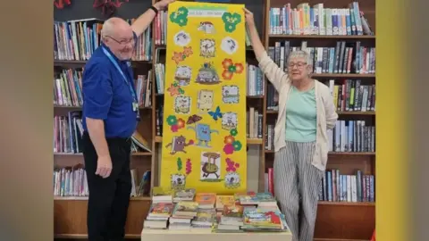 Friends of Woodston Library Volunteers - a man wearing a blue T-shirt and black trousers and a woman wearing a mint coloured T-shirt, a white cardigan and striped trousers, holding a yellow poster placed behind a table of books. 