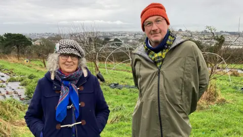 Sarah Pethybridge and Mike Westley stand in a field overlooking Falmouth. Both are wearing coats and hats. They both have scarfs on.