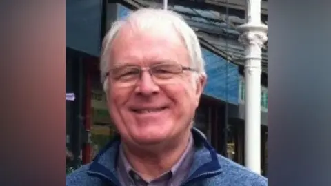 Mike Bath Mike Bath has white hair, spectacles and a grey shirt, with blue kagoule and stands outside the Etihad Stadium. It is a head and shoulders shot.
