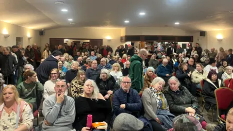 Local Democracy Reporting Service A room full of people sitting in rows and standing around the perimeter of a room, facing the camera, waiting for a meeting to start. 