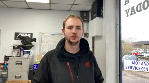 Man stood in car garage. He's wearing a black coat stood by the window. There's an MOT sign and old cabinets full of MOT folders in the background