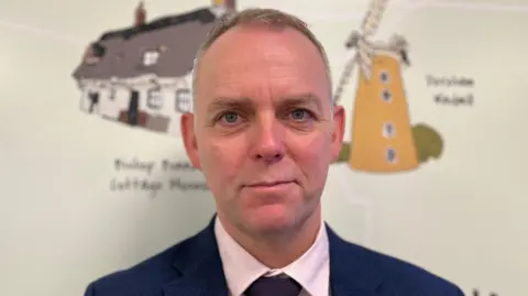 Aimee Dexter/BBC Tristan Ashby is stood in the middle of the image. He has short brown hair and is wearing a pink shirt and a navy tie and suit jacket. Behind him is a mural of a house and windmill. 