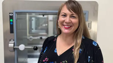 The lab's director, Sarah Albon. She has long blonde highlighted hair and is wearing pink lipstick and a black floral dress. She is smiling into the camera. Behind her is a piece of equipment.