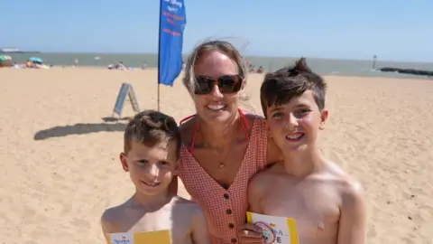 Two young boys stand either side of their mum. She is wearing a red gingham, buttn down dress and is wearing sunglasses .They are standing on a sany beach with a flag and people playing in the background.