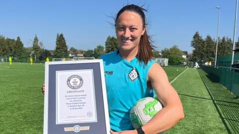 Megan is smiling at the camera holding a football in her left arm and her Guinness World Record certificate in her right arm.  She has brown hair tied back in a pony tail. The certificate is white rimmed and navy.  Her football kit is baby blue with a white triangular shaped lion badge. Behind her is a green astro turf pitch with white goals and yellow mannequins visible in the distance.  