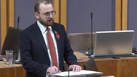 Senedd Cymru Jack Sargeant speaking in the Senedd chamber with his hands rested on papers on top of a lectern. He has a beard and and moustache and is wearing a dark grey suit with a poppy on his lapel, a white shirt and a tie. There are two microphones pointed upwards on the empty desks behind and the silver coloured backs of computer screens.