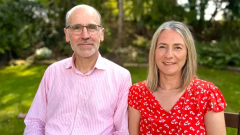  Martin Giles/BBC Guy and Claire sat next to each other outside, smiling into the camera.