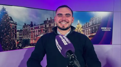 Jack Percival Jack sits in a purple BBC Radio London studio, smiling at the camera with a mic in front of him. A picture of a London street is shown as the background. 