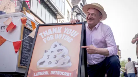 Man wearing straw boater, pink shirt and blue chinos crouched low pointing at a A-board sign reading "Thank you for voting Liberal Democrat".