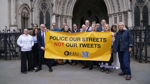 PA Media A group of people standing outside the Royal Courts of Justice holding a yellow banner with the slogan "police our streets not our tweets" written in capital letters. The words are mostly black expect for "not", which is in red. The banner includes the Free Speech Union logo, which is a fist clutching a sharpened pencil and the letters FSU. There is also a black and white QR code on the banner.