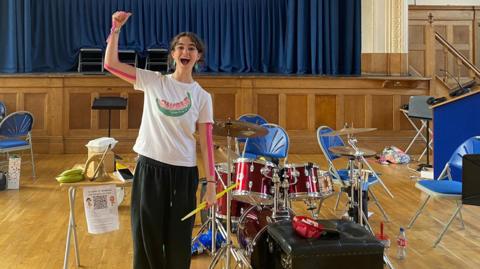 Aev holding her fist aloft, cheering, standing next to her drumkit in the middle of an assembly hall. The stage is behind, with a blue curtain drawn.