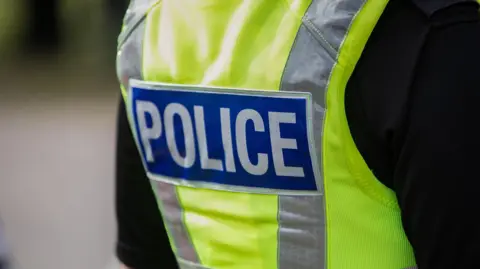 The back of a person wearing police uniform. They are wearing a black t-shirt with a yellow hi-visibility vest featuring a police logo.