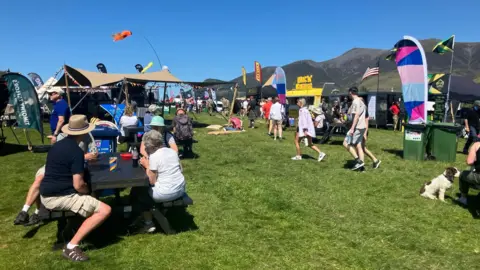 Festival goers checking out the stalls at Keswick Mountain Festival. A group of them is sat around a black wooden table. There are colourful flags and banners on the grassland, with Lake District peaks rising against the clear blue sky.