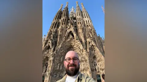 A man with a brown beard in glasses stands in front of a cathedral in Barcelona.