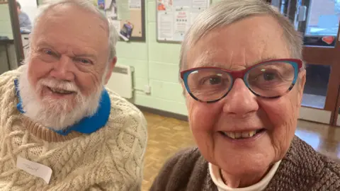 Tony and Wendy smiling to the camera, both sitting in the village hall and wearing woolly jumpers. Wendy has blue and red glasses.