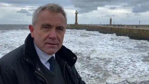 Yorkshire Water A man, Robert Goodwill, wearing a tie looks at the camera with a neutral expression on his face. He is at the coast with the sea and a pier behind him.