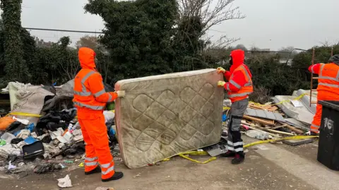 Two men in orange hi viz carry a dirty and stained mattress into a truck