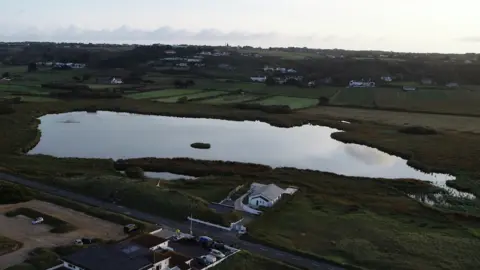 An aerial view of St Ouen's Pond surrounded by fields behind it with a few houses and in front a main road with a car park on the other side and another building. Sunset skies.