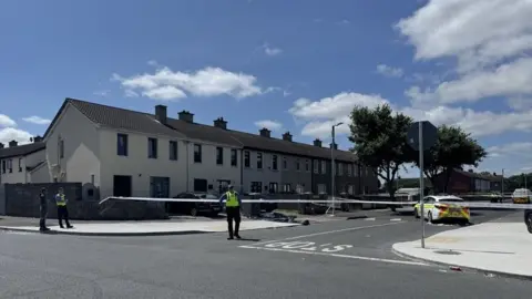RTÉ Scene in Shancastle, west Dublin, after a fatal assault. There are a number of houses, a marked garda car behind tape and officers in yellow high visibility jackets in front of the tape.