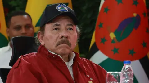 President of Nicaragua Daniel Ortega looks on during the opening event of the XXIII Summit of the Bolivarian Alliance for the Americasin Caracas, Venezuela, in 2024. He is wearing a red jacket and a baseball cap with the Nicaraguan flag emblazoned on it. He has a moustache. 