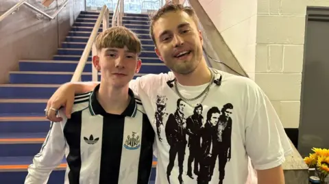 Joe Bartley and Sam Fender back stage at St James' Park. Fender has his arm around Joe who is wearing a Newcastle United shirt.