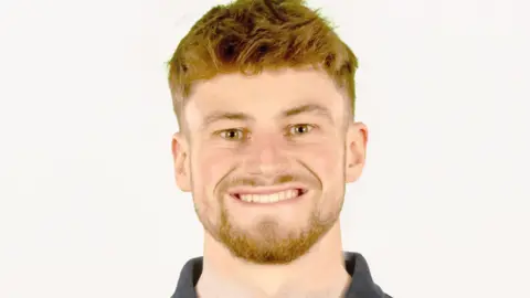 Joe Shingler, wearing a Team UK grey polo shirt. He has short ginger hair and beard and is smiling at the camera against a white backdrop 
