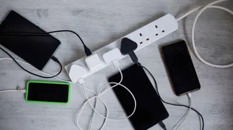 Getty Images/Sally Anscombe Two mobile phones and two tablet computers lying on a grey wood-effect floor next to a white six-socket extension lead.  The devices are each attached to chargers which are plugged into the adapter.  There are two sockets not in use. 