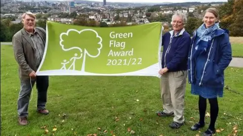 LDRS Two men hold a green flag in a park in Sheffield which has the city skyline in the background. A woman stands next to them on the right. 
