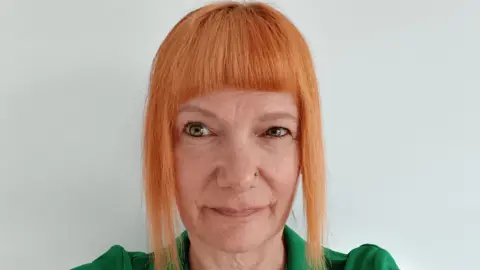 Chrissy has orange hair and is wearing a bright green jacket. She has pale green eyes and is staring just off to the right of the camera. She has a small silver nose stud on the left side of her nose.