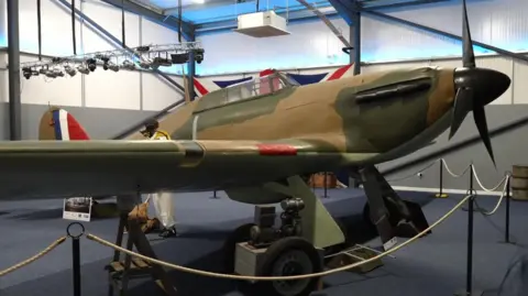 A full-size replica Hurricane plane. The camera is positioned to the front left, looking down the wing towards the fuselage. The plane is painted in a green and brown camouflage scheme. with a black propeller and nose cone. RAF tail markings, in blue, red and white can be seen. A mannequin dressed as a pilot stands behind the wing. The plane is housed in a large, modern warehouse-style building, with a large bank of spotlights hanging from the roof. Ropes cordon off the plane.