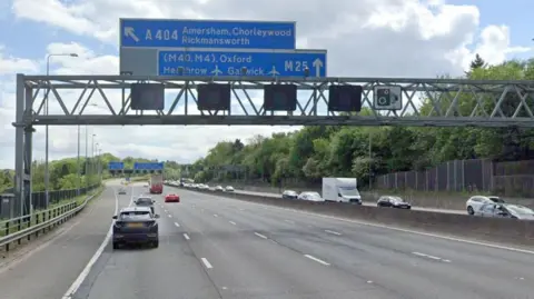 Blue M25 sign on a gantry over the road, show the exit for junction 18 Rickmansworth. There are some cars on the road in both directions.