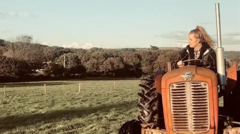 Milly Collins A woman on a farm driving a tractor on a sunny day. She is looking away from the camera.