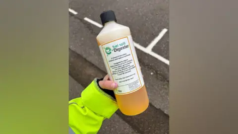 Essex County Council A hand holding a bottle of "fuel spill Digester" - an orange-coloured liqued in a white plastic bottle with a label on it.