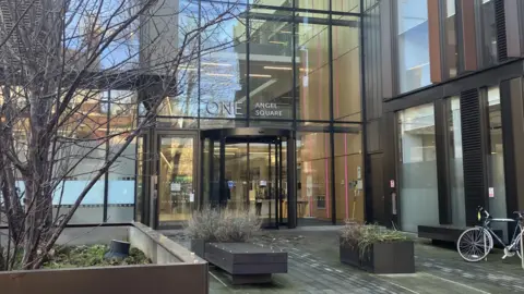 Nadia Lincoln/LDRS The entrance to West Northamptonshire Council's headquarters. It is a large building made up of clear glass panels. A revolving door can be seen as well as some benches, a bike and planted areas.