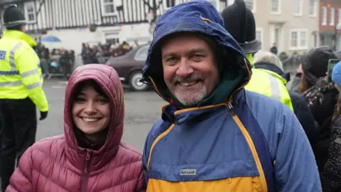 John Fairhall/BBC (L-R) A woman wearing a purple raincoat smiling at the camera and standing next to her dad who is wearing a blue and yellow coat. He is also smiling.