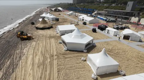 Jamie Niblock/BBC Large tents are erected on a beach. People work on the site and there is some heavy machinery to help manoeuvre equipment.