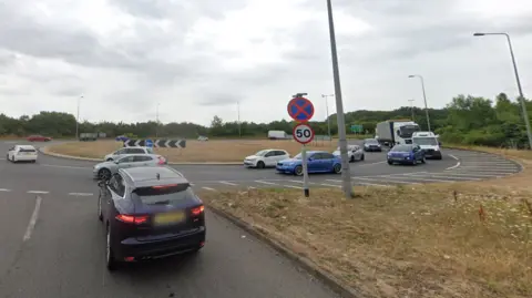 Google A two-lane approach to a roundabout. There are cars on the roundabout and a 50mph road sign.