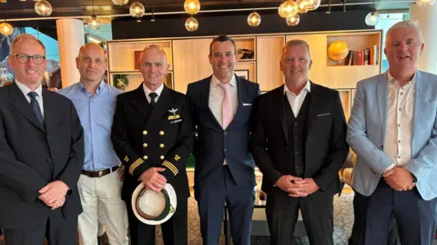 Paraic Slattery The image shows six men standing side by side facing the camera. They are all dressed in suits, bar one who is in a uniform. 