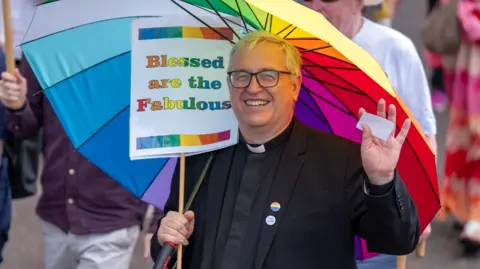 PA Media A participant in a Pride march stands beneath a large rainbow-coloured umbrella while holding a sign reading “Blessed are the Fabulous.” The participant wears a clerical collar and dark jacket and waves toward the camera.