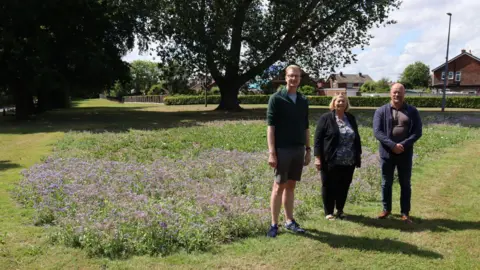 From left, Stockton councillors Niall Innes, Lynn Hall and Jason French on land at the junction of Birkdale Road and Darlington Road, Hartburn, Stockton, where they have planted wildflowers. They are standing on grass with a small patch of wildflowers behind them and trees. It is a sunny day.