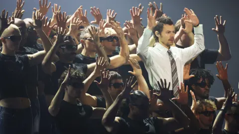 Reuters Harry Styles on stage in the midst of numerous dancers, all holding both hands in the air