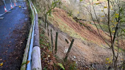 Westmorland and Furness Council A picture taken from the road looking down at the landslip. Fence posts are dangling in the air above a track of brown, wet soil.