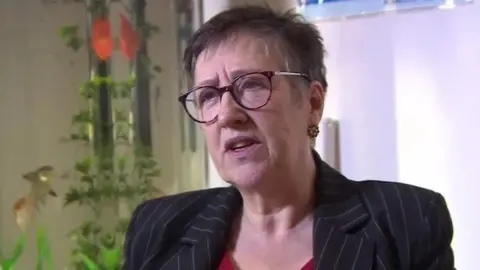 Dr Leigh Vallance speaks in a TV interview wearing a red top and black striped blazer with a fish tank seen in the background.