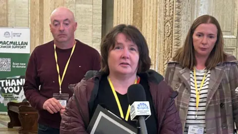 Noeleen Mullan speaking at a microphone. She has shouldel-length brown hair, wearing a burgundy raincoat and black top.
